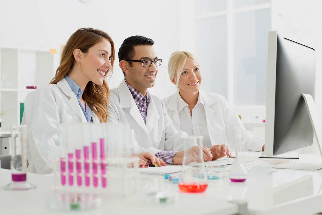 Trzech uśmiechniętych naukowców w białych fartuchach laboratoryjnych, zebranych za ekranem komputera